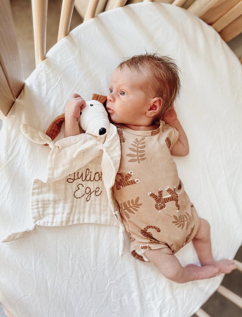 May include: A baby in a brown and white onesie with a tiger print lies in a crib. The onesie has brown leaves and tigers printed on it. The baby is holding a white and brown stuffed animal with the name "Julia Ege" embroidered on a white muslin blanket.