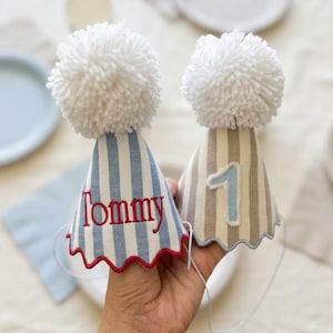 Può includere: Due cappellini da festa conici con pon pon di lana bianca sulla punta. Uno presenta righe verticali blu e bianche con il nome Tommy ricamato in rosso. L'altro ha righe verticali beige e bianche con un'applicazione del numero 1 in azzurro.