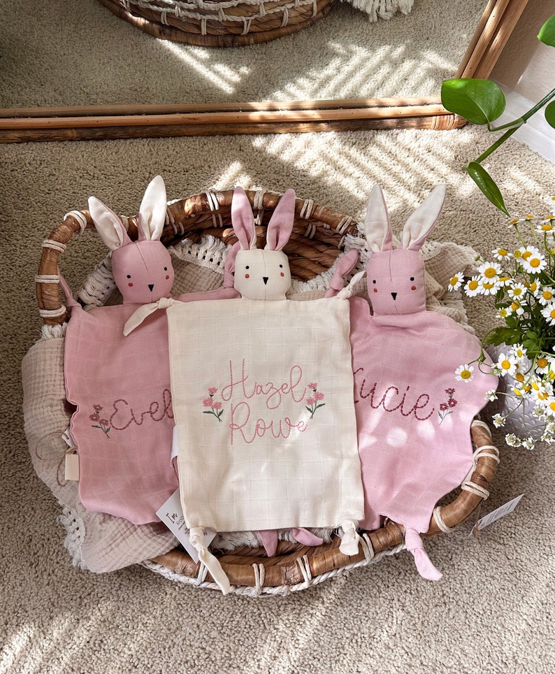May include: Three pink bunny lovey security blankets with embroidered names "Evel", "Hazel Rowe", and "Lucie". Each bunny has a white face and long ears. The blankets are in a woven basket.