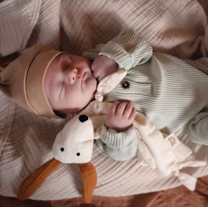 May include: A baby wearing a light green ribbed onesie and a brown knit hat is sleeping with a white and brown stuffed animal. The baby is holding the stuffed animal in their hand.