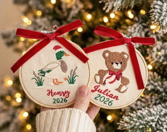 May include: Two round wooden embroidery hoop ornaments featuring cream fabric. One displays a mallard duck in a Santa hat with the text Henry 2026. The other shows a brown teddy bear with a red bow and the text Julian 2026. Both have red velvet bows.