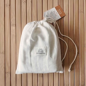 May include: A white drawstring bag with a textured grid pattern, featuring a small rectangular label with the "minivalley" logo. The bag is made of a soft, light-colored fabric and is tied with a white rope. A product tag hangs from the drawstring.