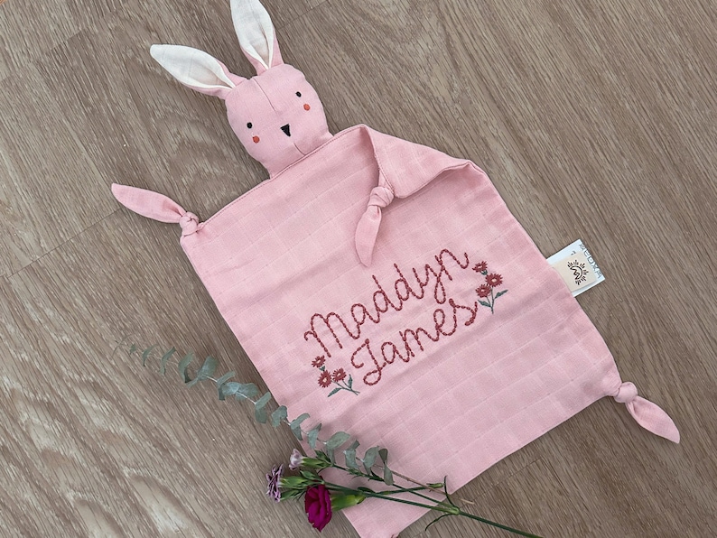 Pu&ograve; includere: Coperta di sicurezza rosa a forma di coniglio con il nome "Maddyn James" ricamato in rosso con piccoli fiori rossi.