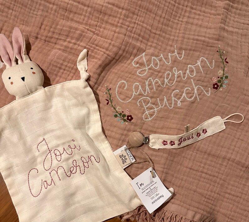 Pu&ograve; includere: Una copertina per neonati in mussola rosa e bianca con il nome "Joui Cameron Busch" ricamato in filo rosa. &Egrave; incluso un bavaglino di coniglio bianco con lo stesso nome ricamato. &Egrave; incluso anche un fermasucchiotto rosa e bianco con il nome "Joui" ricamato.