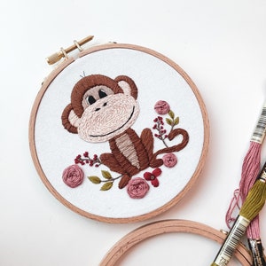 May include: Embroidered hoop art featuring a brown monkey with pink and red flowers. The monkey is sitting on a branch with leaves and flowers. The hoop is made of wood and has a gold screw.