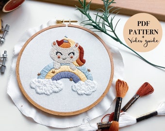 May include: A white embroidery hoop with a colorful embroidered design of a unicorn sitting on a rainbow with clouds. The text "PDF PATTERN + Video guide" is on a peach-colored circle in the upper right corner of the image.
