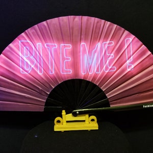 May include: A pink and white folding fan with the words "BITE ME!" printed in white on the front. The fan is made of silk and has a black handle. The fan is sitting on a yellow stand.