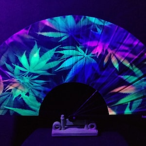 May include: A black folding fan with a colorful marijuana leaf design. The fan is made of paper and has a black handle. The fan is glowing under a black light.