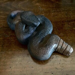 May include: A dark grey, textured sculpture of a snake coiled in on itself. The snake's head is raised, and its body has a rough, dotted texture. The tail end has a segmented design.