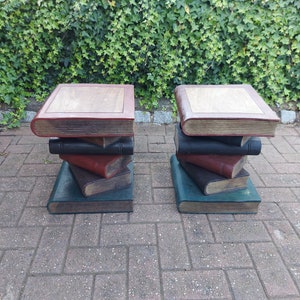 May include: Two wooden book-shaped stools with a stacked book design. The stools are made of wood and painted in shades of brown, red, and green. The stools are sitting on a brick patio.
