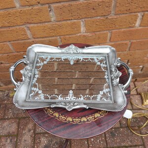 May include: A rectangular silver-toned serving tray with ornate, raised scrollwork detailing along the edges and handles. The centre features a clear glass panel decorated with a delicate, white, lace-like filigree pattern around the perimeter.