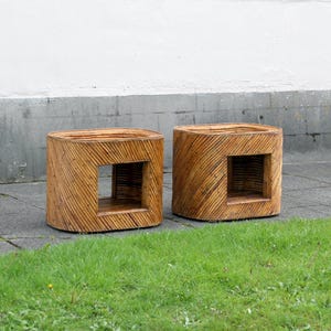May include: Two square, wooden side tables with a natural finish. Each table has a rectangular opening on one side and is constructed from thin, diagonal wooden slats. The tables are placed on a concrete surface with green grass in the foreground.