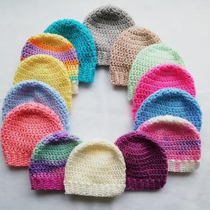 May include: A collection of colorful, handmade crochet beanies arranged in a circular pattern. The beanies showcase a variety of colors, including pink, blue, yellow, and purple, with some featuring striped patterns. The hats are made of soft yarn.