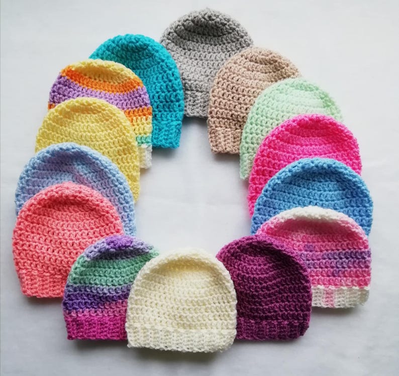 May include: A collection of colorful, handmade crochet beanies arranged in a circular pattern. The beanies showcase a variety of colors, including pink, blue, yellow, and purple, with some featuring striped patterns. The hats are made of soft yarn.