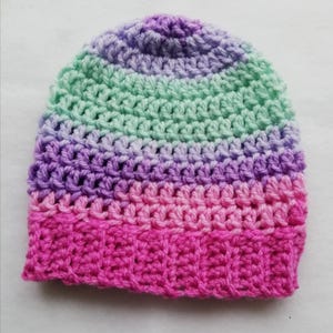 May include: A handmade crochet beanie in a gradient of colors, transitioning from pink to purple and ending in a light green hue. The hat is made with a textured stitch, showcasing the craftsmanship of the item.
