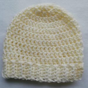 May include: A cream-colored, crocheted beanie hat. The hat is made with a textured stitch pattern and features a ribbed brim. The hat is a solid color and appears to be handmade.