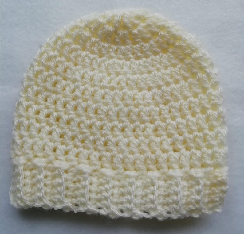 May include: A cream-colored, crocheted beanie hat. The hat is made with a textured stitch pattern and features a ribbed brim. The hat is a solid color and appears to be handmade.