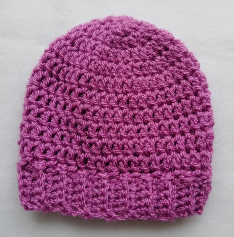May include: A handmade, magenta-colored crochet beanie hat. The hat features a textured stitch pattern throughout the crown and a ribbed brim. This warm hat is perfect for cold weather and is a stylish accessory.
