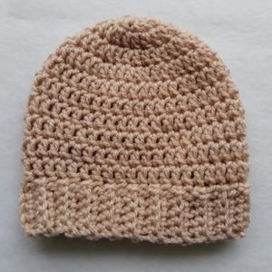 May include: A crocheted beanie hat in a light tan color. The hat features a ribbed brim and a dense, textured stitch pattern throughout the crown. The hat is made of a soft, warm yarn, perfect for cold weather.