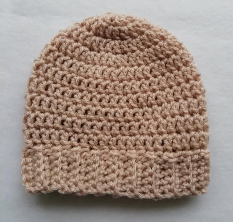 May include: A crocheted beanie hat in a light tan color. The hat features a ribbed brim and a dense, textured stitch pattern throughout the crown. The hat is made of a soft, warm yarn, perfect for cold weather.