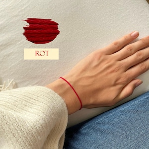 Könnte beinhalten: Ein rotes Armband aus Faden wird am Handgelenk getragen. Das Wort "ROT" wird in einem cremefarbenen Rechteck angezeigt. Das Armband ist eine einfache, dünne Schnur, und die Farbmuster über dem Text ist ein leuchtendes Rot.