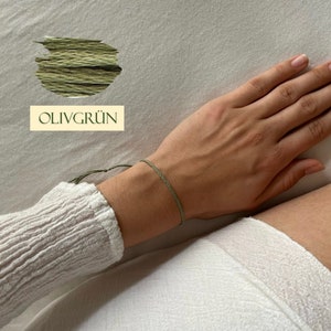 Könnte beinhalten: Zartes olivgrünes Armband am Handgelenk. Das Bild zeigt eine Probe olivgrünen Fadens und den Text "OLIVGRÜN". Das Armband ist ein schlichtes, minimalistisches Design, das sich für den Alltag eignet.