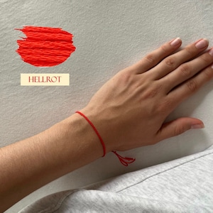 Könnte beinhalten: Ein rotes Armband aus Faden wird am Handgelenk getragen. Das Wort "HELLROT" wird in einem rechteckigen Feld angezeigt. Ein roter Pinselstrich befindet sich in der oberen linken Ecke.