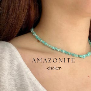 Op de afbeelding: Een delicate amazoniet choker ketting met kleine, onregelmatige turquoise kralen. De ketting wordt om de nek van een persoon getoond. Het woord "AMAZONITE" en "-choker-" zijn in zwarte tekst eronder gedrukt.