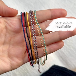 Peut inclure: Une collection de bracelets en cordon torsadé fin dans divers motifs unis et multicolores, notamment rouge, bleu marine, noir, jaune et sarcelle. Une incrustation graphique blanche affiche le texte 70+ colors available.