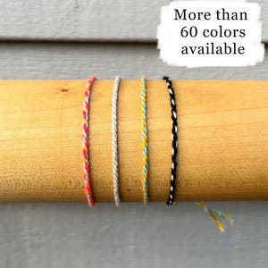 Può includere: Quattro braccialetti dell'amicizia intrecciati in colori vivaci: rosa, bianco, giallo e nero. I braccialetti sono esposti su un tassello di legno su sfondo grigio. Il testo nell'immagine recita: "Più di 60 colori disponibili."