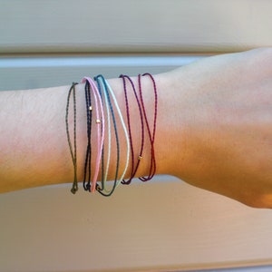 Könnte beinhalten: Eine Sammlung dünner Armbänder aus Schnur in verschiedenen Farben, darunter Grün, Schwarz, Rosa und Burgunderrot. Jedes Armband hat eine kleine goldene Perle. Die Armbänder werden an einem Handgelenk gezeigt.
