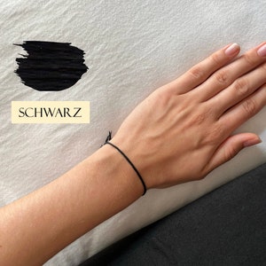 Könnte beinhalten: Ein minimalistisches Armband aus schwarzem Faden wird an einem Handgelenk gezeigt. Das Wort "SCHWARZ" befindet sich in einem rechteckigen Feld im Bild. Der Hintergrund ist ein weißes Laken.