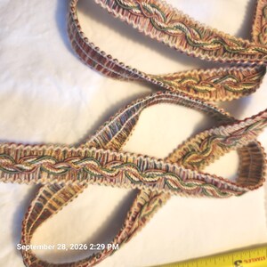 May include: A decorative gimp trim featuring a braided pattern in shades of green, red, gold, and cream. The trim has a scalloped edge and is positioned next to a yellow measuring tape showing a portion of the tape measure in inches.