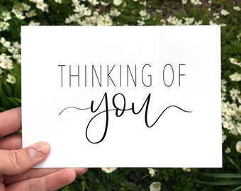May include: A white card with the words "THINKING OF YOU" in black script. The card is held in front of a background of green foliage and white flowers. The text is in a simple, elegant font.