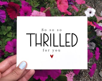 May include: A white card with the words "So so so THRILLED for you" in black lettering and a small red heart. The card is held in a hand with light blue nail polish, set against a backdrop of vibrant pink and purple flowers.