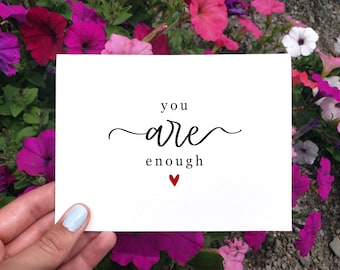 May include: A white card with black text that reads "you are enough" with a small red heart below the text. The card is held by a hand against a background of pink and white flowers.