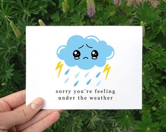 May include: A white card with a blue cartoon cloud with a sad face and raindrops. The text on the card reads "sorry you're feeling under the weather".