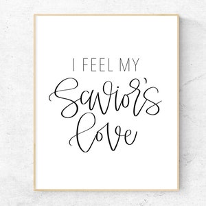 May include: A white framed print with black calligraphy that reads "I feel my Savior's love".
