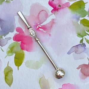 Peut inclure: Infuseur à thé en argent avec une boule ronde perforée à l'extrémité et un manche décoratif avec un motif floral. L'infuseur est sur un fond blanc avec des fleurs roses et vertes en aquarelle.
