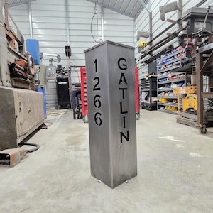 May include: A tall, rectangular metal address sign with the numbers "1266" and the name "GATLIN" cut out. The sign is a muted gray color and stands on a concrete floor in a workshop setting.