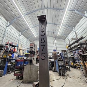 May include: A tall, dark metal address post with the numbers 53875 cut out. The post has a small, square top and is set in a workshop with various tools and equipment. The numbers are large and easy to read.