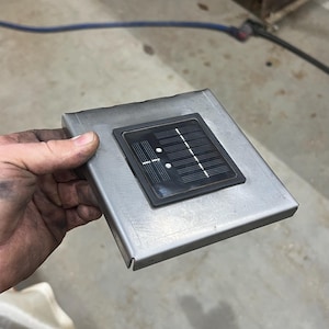 May include: A square, brushed metal component with a black solar panel inset. The solar panel has a grid pattern and two white dots. The metal component is held in a hand.