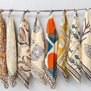 May include: Ten rectangular fabric cloths hanging from a horizontal rope. Each cloth features a unique printed pattern, including floral motifs, paisley, leopard faces, geometric shapes, fish, and a green and white checkerboard design on cream fabric.