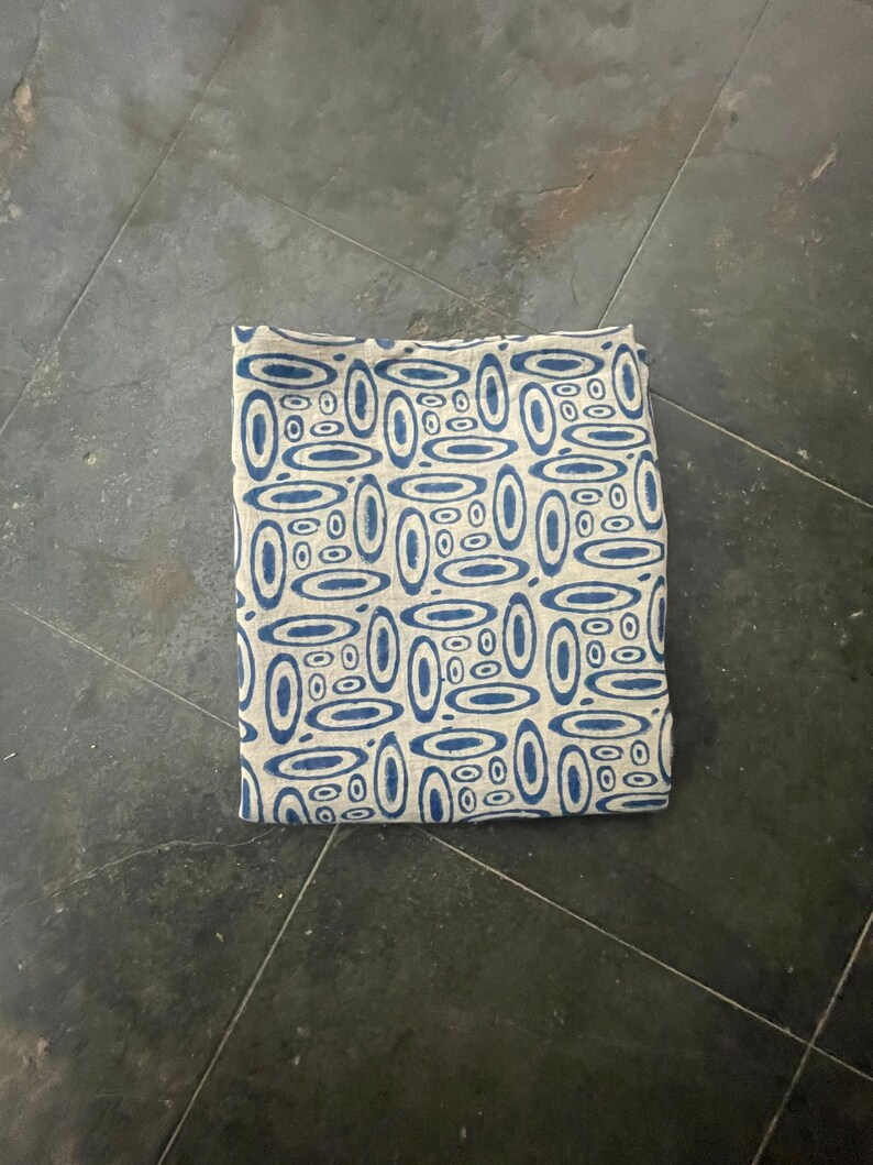 May include: A blue and white patterned fabric with a geometric design of overlapping circles. The fabric is folded in half and lying on a gray surface.