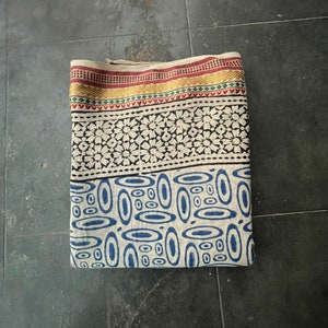 May include: A folded fabric with a white background and blue, black, red, and gold patterns. The fabric has a border with a geometric design.