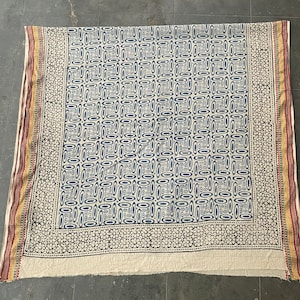 May include: A white cotton fabric with a blue and white geometric pattern. The fabric has a border with a black and white floral design. The fabric is folded in half and has a frayed edge.