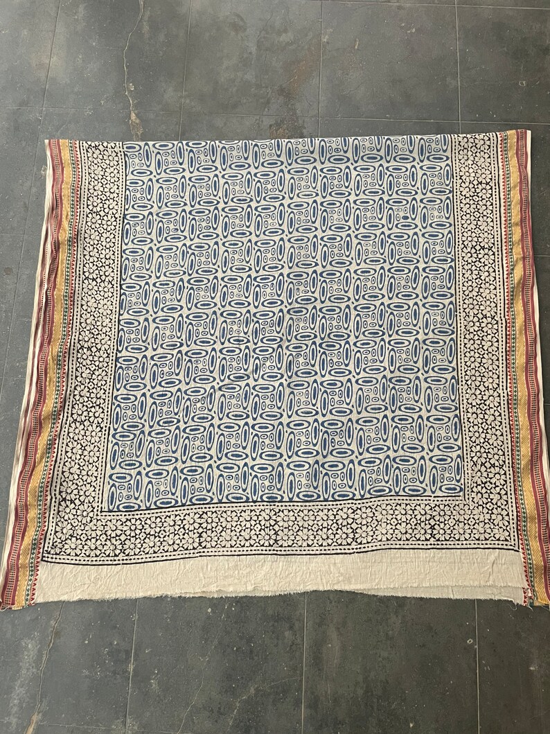 May include: A white cotton fabric with a blue and white geometric pattern. The fabric has a border with a black and white floral design. The fabric is folded in half and has a frayed edge.