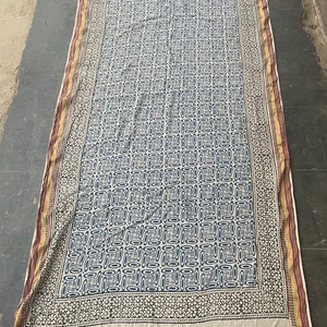 May include: A white cotton fabric with a blue and black geometric pattern. The fabric has a border with a floral design in black and white.