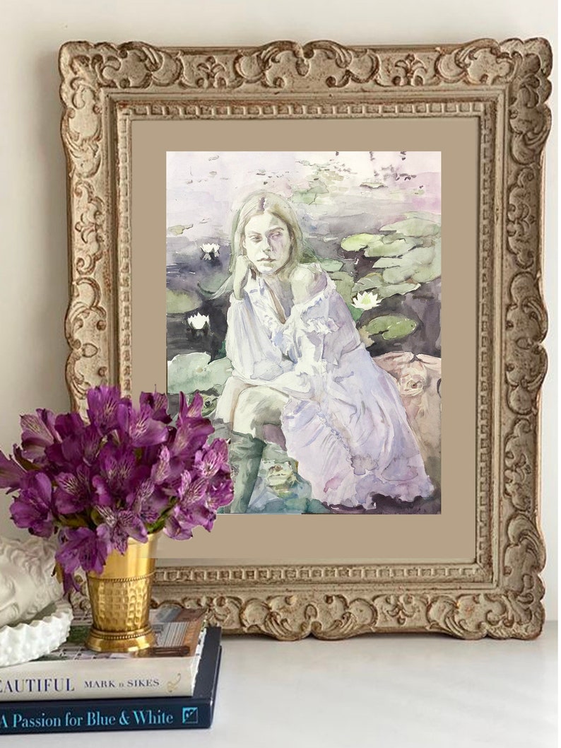 May include: A framed watercolor painting depicts a person in a white dress seated near water lilies. The painting is in a decorative, ornate, gold-toned frame. A bouquet of purple flowers in a gold vase and books are in the foreground.