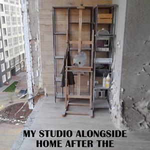 May include: An artist's studio on a balcony, featuring a wooden easel, a metal shelving unit, and a birdcage. The text "MY STUDIO ALONGSIDE HOME AFTER THE RUSSIAN INVASION IN BUCHA" is visible. The balcony shows signs of damage.